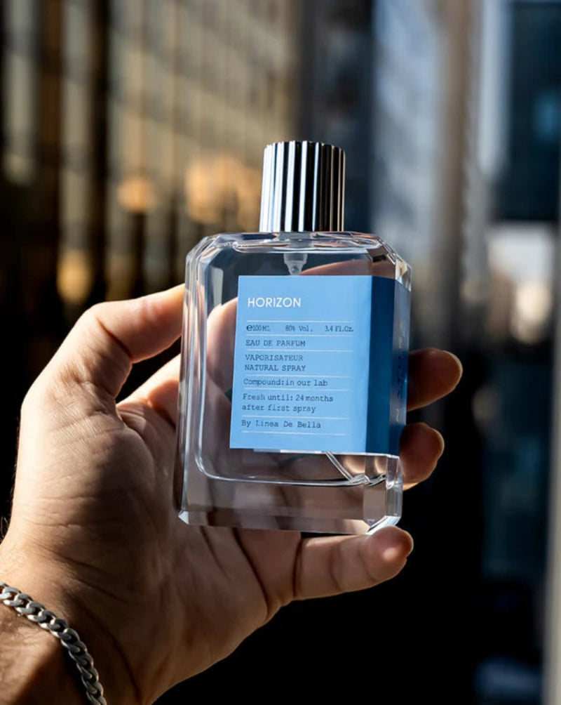 Hand holding a clear perfume bottle with a blue label against a blurred cityscape background