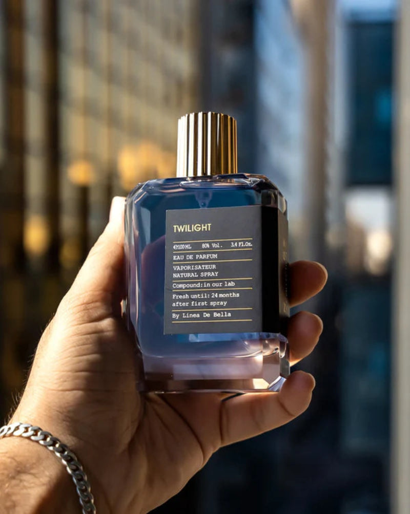 Hand holding a perfume bottle labeled 'Twilight' with a blurred cityscape background